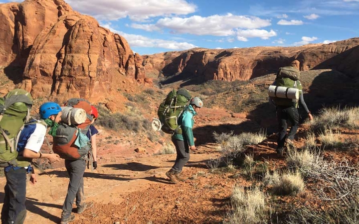 A group of people wearing backpacks hike along a trail through a desert landscape. There are tall canyon walls in the background.